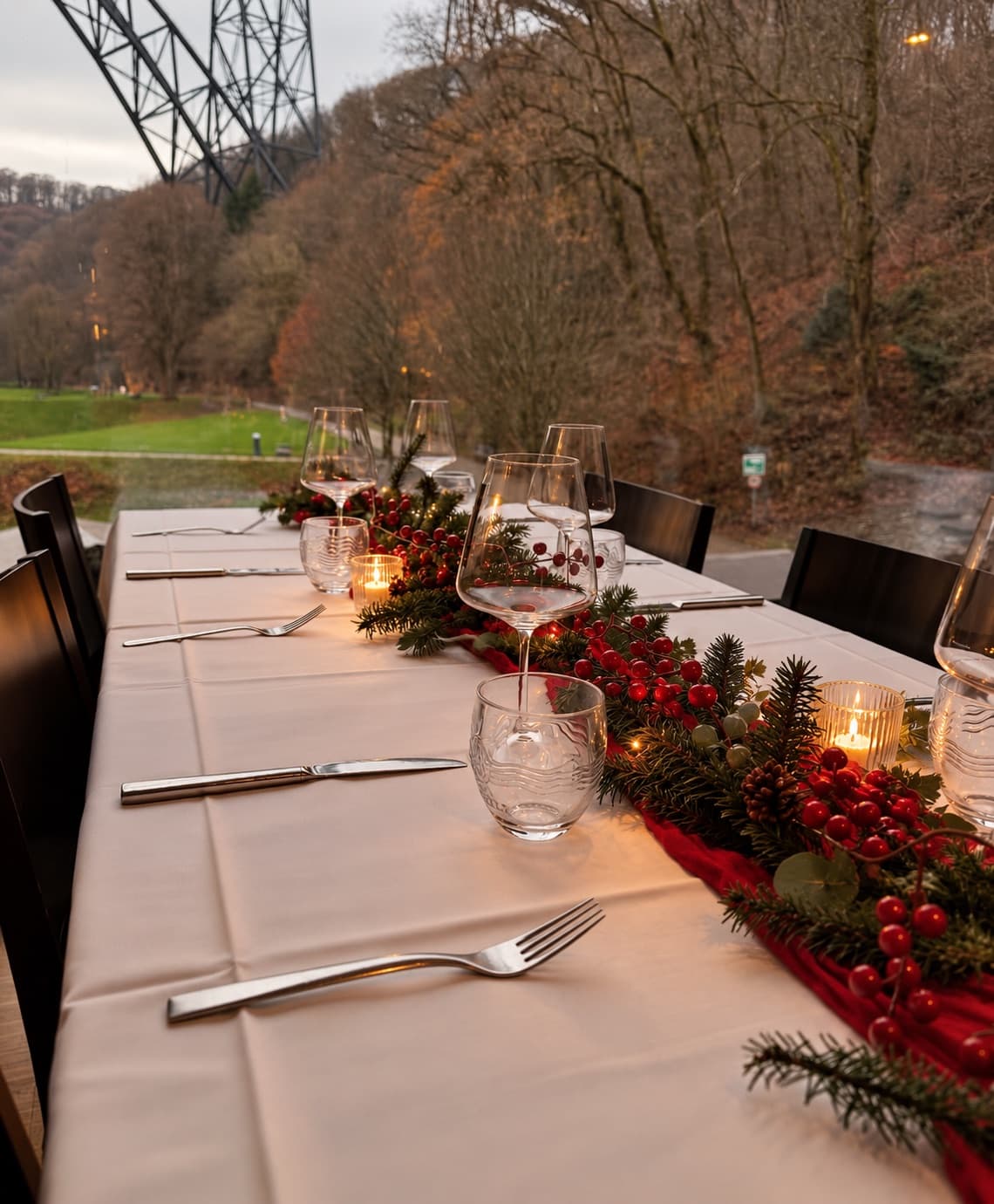 Weihnachtlich gedeckte lange Tafel am Fenster mit rotem Läufer, Tannengrün, roten Beeren und Kerzen, draußen das winterliche Tal und ein Pfeiler der Müngstener Brücke
