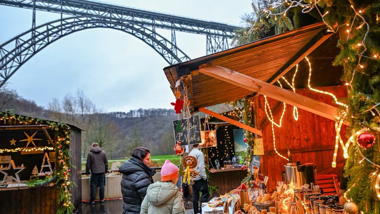 Beleuchtete Holzhütten mit Tannengrün und Lichterketten beim Winterdorf, dahinter der Bogen der Müngstener Brücke in der Dämmerung